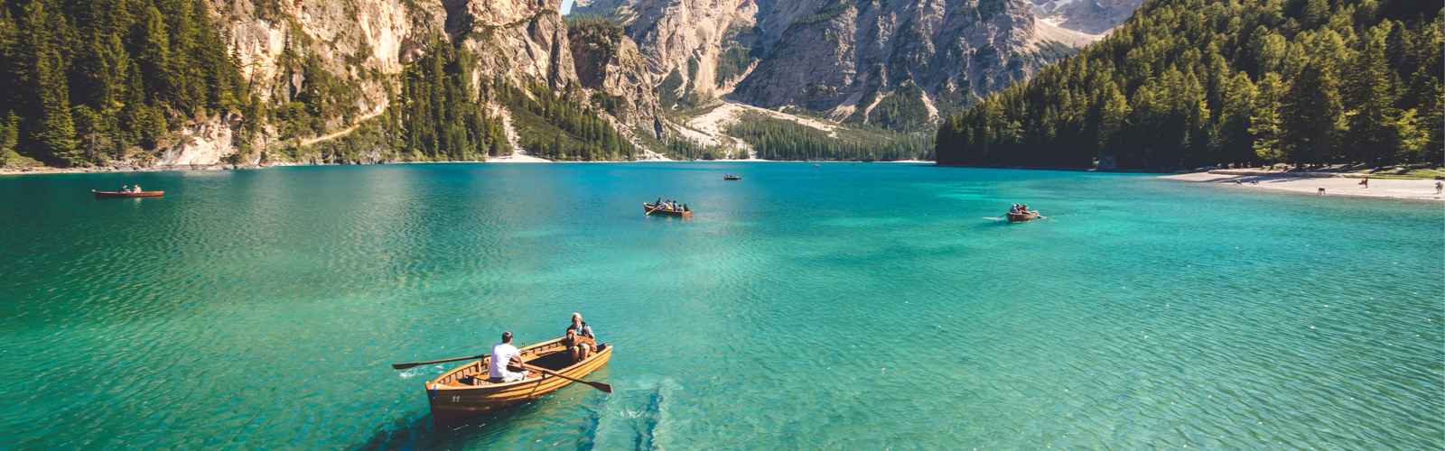 A couple and their dog sitting in their rowboat over crystal clear waters, with mountains and forests in the background.