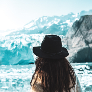 A woman with long brown hair and a black hat looking towards some snowy mountains.